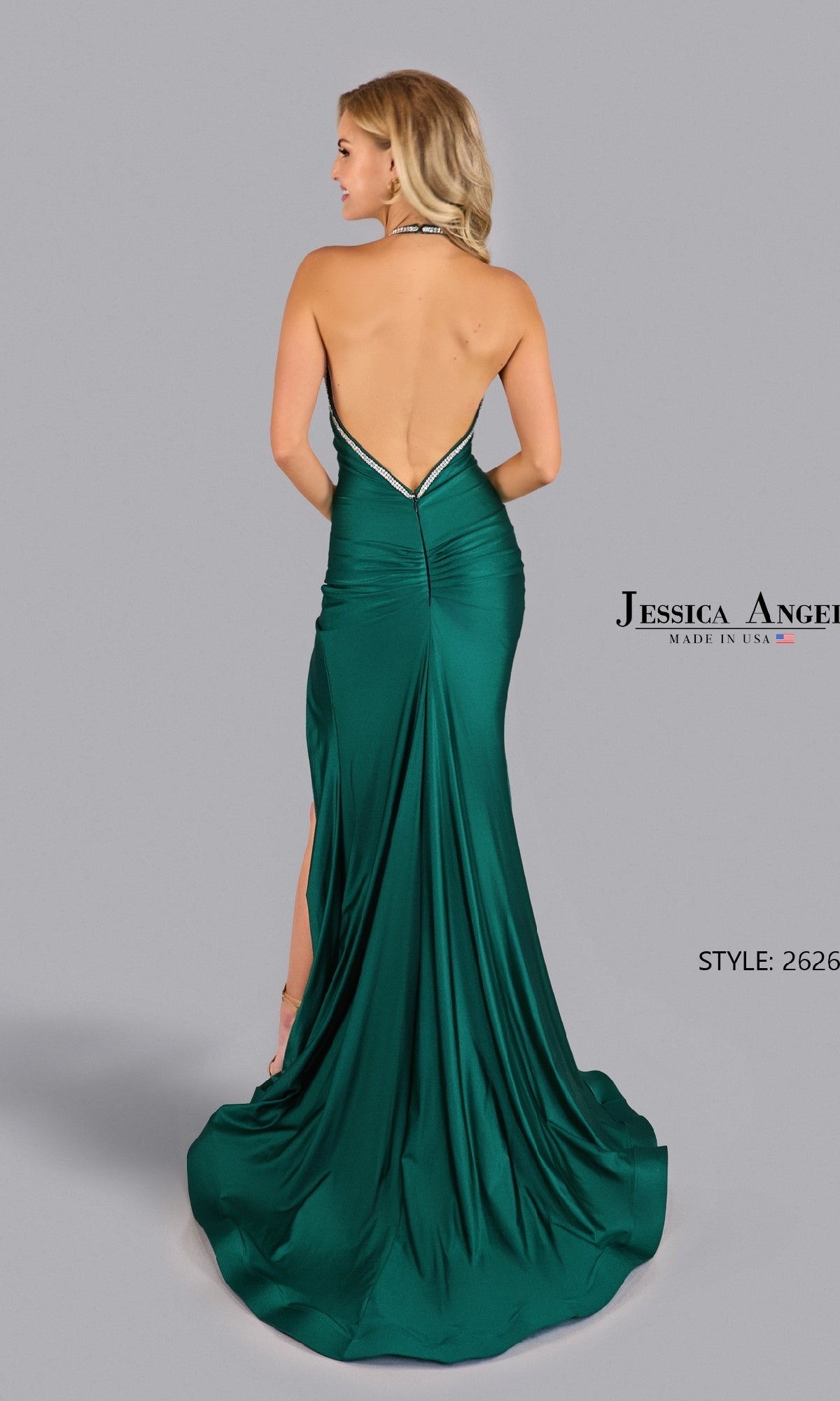 A woman models the Jessica Angel 2626 Open-Back Halter Prom Gown in emerald green, featuring a backless design and long train, posing with her back to the camera against a plain gray background.